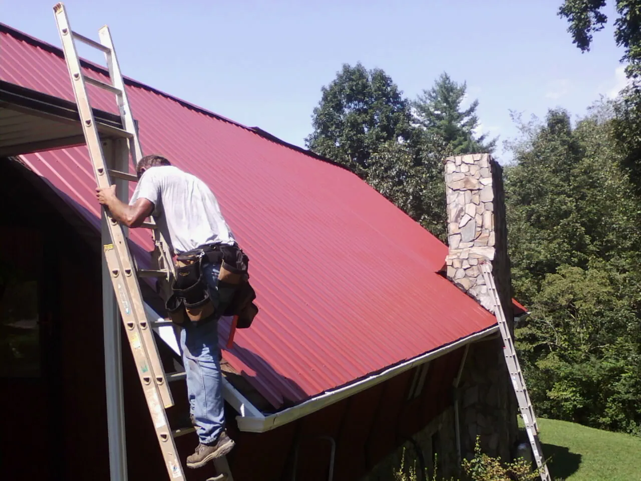 Roofer on a ladder beside a red metal roof and stone chimney — Soffit & Fascia Repair by Roofing Brothers in Saint Henry